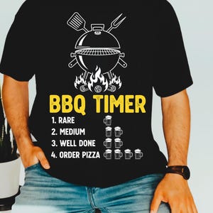 May include: Black t-shirt with a white graphic of a barbecue grill, spatula, and fork. The text "BBQ TIMER" is in yellow, with cooking levels and beer mug icons below. The shirt is worn by a person.