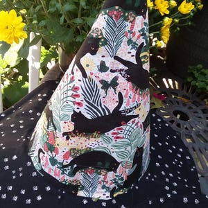 May include: A black witch hat with a white, pink, and green floral pattern featuring black cat silhouettes and butterflies. The brim is black with white paw prints. The hat is cone-shaped and sits on a black surface.