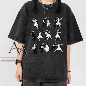 Puede incluir: Una camiseta gris carbón con efecto lavado al ácido que presenta un estampado gráfico de nueve parejas de bailarines en blanco y negro. El diseño muestra varias poses de baile de salón, incluyendo giros y caídas, dispuestas en una cuadrícula.