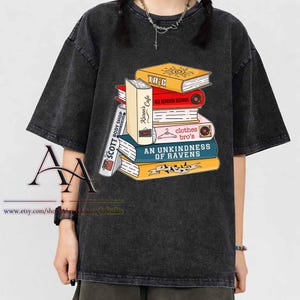 May include: A charcoal gray acid-wash t-shirt featuring a graphic of a stack of colorful books. The book spines display text including "Karen's Cafe," "RED BEDROOM RECORDS," "clothes bro's," "AN UNKINDNESS OF RAVENS," and "SCOTT BODY SHOP."