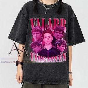 以下が含まれることがあります： 男性のグラフィックとピンク色の「VALARR TARGARYEN」の文字が入った濃いグレーのTシャツ。 ヴィンテージ感のある、使い古したような外観です。 EtsyショップのURLがTシャツの下部に表示されています。
