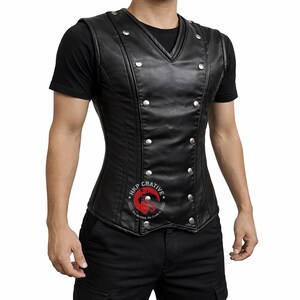 May include: A black leather vest with a V-neck and silver-colored button details. The vest has a double-breasted design and a fitted silhouette. The vest is sleeveless and appears to be worn over a black t-shirt.