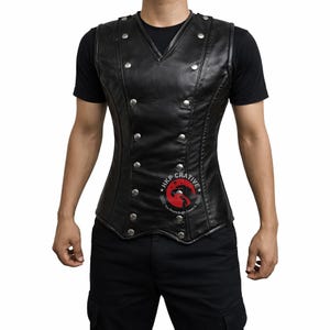 May include: Black leather vest with silver-colored buttons. The vest has a V-neck and a fitted design. The vest is worn over a black t-shirt. The vest has a logo on the lower right side.