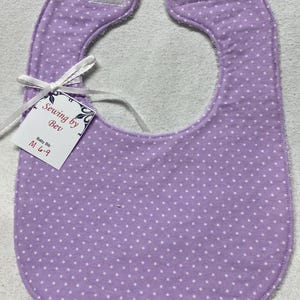Może przedstawiać: Fioletowy śliniak dla niemowlęcia w białe kropki z białą wstążką. Śliniak ma zapięcie na rzep i metkę z napisem "Sewing by Bev" i "Baby Bib M 6-9".