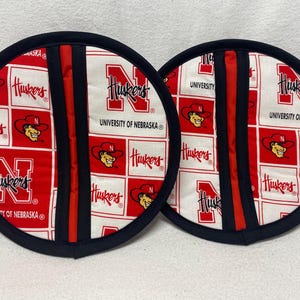 May include: Two red and white pot holders with a black trim. The pot holders feature the University of Nebraska Huskers logo and the words "Huskers" and "University of Nebraska".