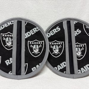 May include: Two round pot holders featuring the Raiders logo in black and white. The pot holders have a black background with the Raiders logo and the word "RAIDERS" printed on them. The edges are gray.
