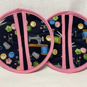 May include: Two pink and black pot holders with a sewing theme. The pot holders feature a black background with colorful sewing-themed designs, including buttons, thread spools, needles, and a sewing machine.