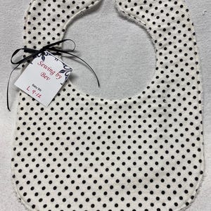 Może przedstawiać: Biały śliniak dla niemowlęcia w czarne kropki. Śliniak ma strzępiony brzeg i jest związany czarną wstążką. Do wstążki przymocowana jest metka z napisem "Sewing by Bev" i "Baby Bib L 9-12".