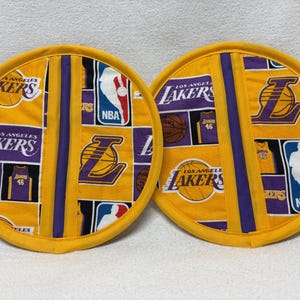 May include: Two yellow and purple pot holders with a Los Angeles Lakers theme. The pot holders feature a basketball, jersey, and the team logo.