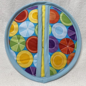 May include: A blue pot holder with a colorful geometric pattern of circles and stripes. The pot holder is made of fabric and has a yellow trim.