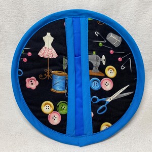 Może przedstawiać: Okrągła, niebiesko-czarna rękawica do garnków z motywem szycia. Rękawica do garnków przedstawia maszynę do szycia, guziki, nici, igły i manekin.