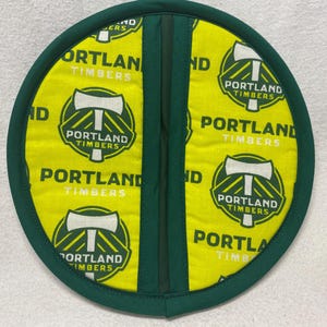 May include: A green and yellow round pot holder with the Portland Timbers logo repeated on the fabric. The logo features a green and white axe with the words "Portland Timbers" written in white.