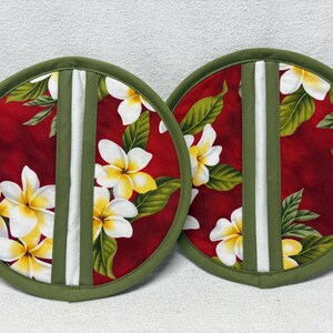 May include: Two round pot holders with a red background featuring white and yellow plumeria flowers and green leaves. The edges are green, with a white and green vertical stripe down the center. The pot holders are made of fabric.