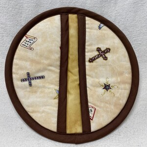 May include: A round brown pot holder with a beige center. The center has a design of crosses, stars, and scrolls. The pot holder has a brown and yellow stripe down the center.