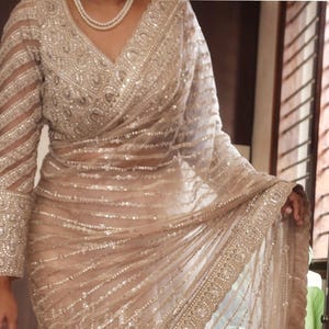 May include: A beige saree featuring intricate silver embroidery, sequins, and beadwork throughout the fabric. The garment includes a long-sleeved blouse with a V-neckline and a matching border with detailed patterns, paired with a pearl necklace.