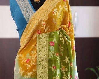 May include: A multi-colored saree featuring a gradient of mustard yellow, olive green, and bright blue. The fabric is adorned with a floral pattern in red, white, and pink, complemented by a wide, intricate silver border with woven motifs.