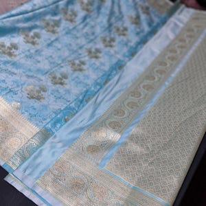 May include: A light blue textile with gold floral patterns and decorative borders. The fabric has a silky sheen and intricate detailing. The borders feature ornate designs, adding a touch of elegance to the overall look. The textile is folded, showcasing its texture and design.