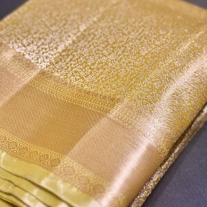 May include: Close-up of a folded, golden-yellow textile with intricate white floral patterns. The fabric has a textured appearance with a contrasting, smooth border. The textile is likely a saree or similar garment.