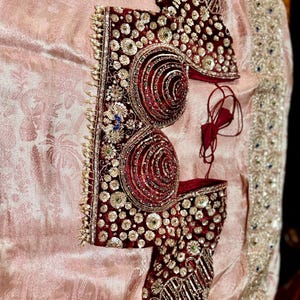 May include: A maroon and gold embroidered garment, possibly a blouse or bodice, with intricate detailing. The garment features a velvet-like texture and is adorned with sequins and beadwork. It rests on a pale pink, patterned fabric.