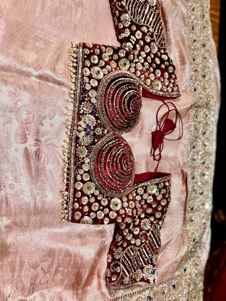 May include: A maroon and gold embroidered garment, possibly a blouse or bodice, with intricate detailing. The garment features a velvet-like texture and is adorned with sequins and beadwork. It rests on a pale pink, patterned fabric.
