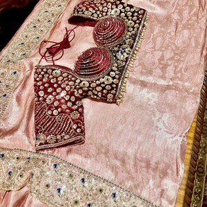 May include: A formal ensemble featuring a light pink fabric with a floral pattern. The set includes a burgundy bodice with intricate beadwork and embellishments. The edges of the fabric are adorned with detailed embroidery and colorful accents.