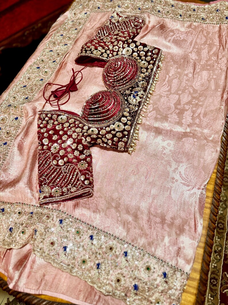 May include: A formal ensemble featuring a light pink fabric with a floral pattern. The set includes a burgundy bodice with intricate beadwork and embellishments. The edges of the fabric are adorned with detailed embroidery and colorful accents.