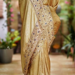 May include: A golden-hued saree with intricate embroidery and sequin detailing. The saree features a decorative border with blue and gold accents. The fabric drapes elegantly, showcasing the design and texture of the garment.