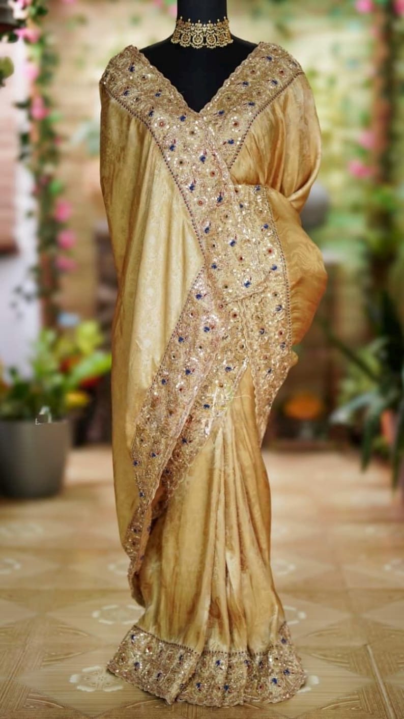 May include: A golden-hued saree with intricate embroidery and sequin detailing. The saree features a decorative border with blue and gold accents. The fabric drapes elegantly, showcasing the design and texture of the garment.