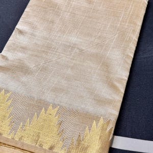 May include: A folded beige fabric textile featuring a subtle horizontal and vertical weave pattern. The bottom edge is adorned with a wide, metallic gold border displaying a repeating chevron or zigzag geometric design across the entire width.