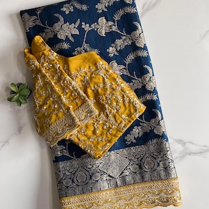 May include: A navy blue saree with a floral pattern and a gold border, paired with a mustard yellow embroidered blouse. The saree is folded neatly, showcasing the intricate design and texture of the fabric.