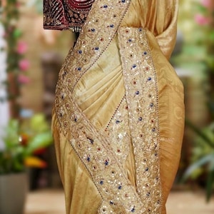 May include: A gold-colored saree with intricate embroidery and embellishments. The saree features a detailed border with sequins and colorful accents. A maroon blouse with ornate embroidery is visible.