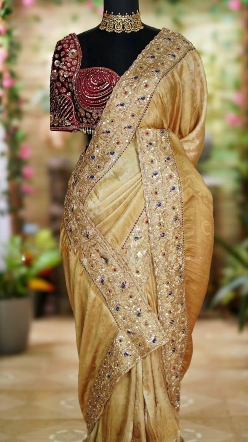 May include: A gold-colored saree with intricate embroidery and embellishments. The saree features a detailed border with sequins and colorful accents. A maroon blouse with ornate embroidery is visible.