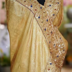 May include: A gold-colored sari with intricate embroidery and embellishments. The sari features a detailed border with blue, red, and white accents. The fabric has a subtle, ornate pattern. A gold-toned necklace is also visible.