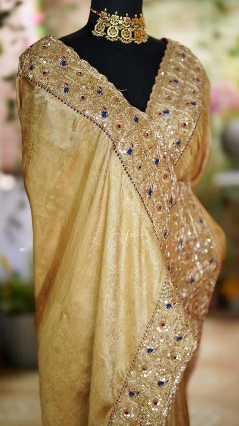 May include: A gold-colored sari with intricate embroidery and embellishments. The sari features a detailed border with blue, red, and white accents. The fabric has a subtle, ornate pattern. A gold-toned necklace is also visible.