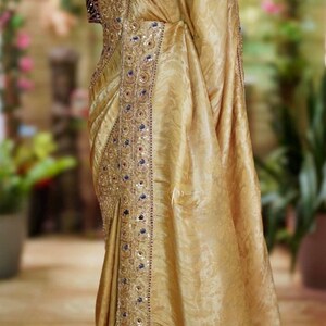 May include: A golden-yellow sari with intricate embroidery and embellishments. The sari features a detailed border with blue and red accents. The blouse is a deep burgundy color. The sari is draped over a mannequin, showcasing its flowing fabric and design.