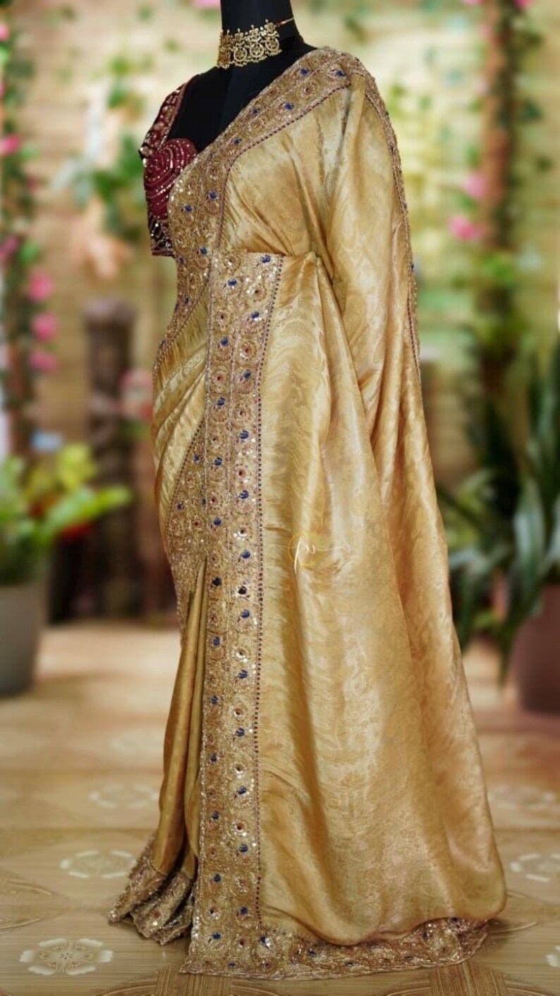 May include: A golden-yellow sari with intricate embroidery and embellishments. The sari features a detailed border with blue and red accents. The blouse is a deep burgundy color. The sari is draped over a mannequin, showcasing its flowing fabric and design.