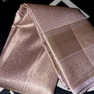 May include: A folded, shimmering, rose-gold textile with intricate patterns. The fabric has a textured appearance and a subtle sheen. The design includes detailed borders with floral and paisley motifs, suggesting a luxurious garment.