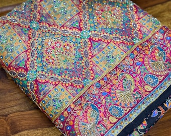 May include: A folded textile featuring a vibrant, multi-colored pattern with intricate embroidery and small reflective embellishments. The design includes geometric shapes, floral motifs, and stylized peacock figures in shades of pink, gold, and blue.