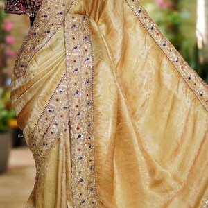 May include: A golden-yellow saree with intricate embroidery and sequin detailing. The border features a pattern of sequins and small embellishments. A maroon blouse with ornate embroidery is visible.