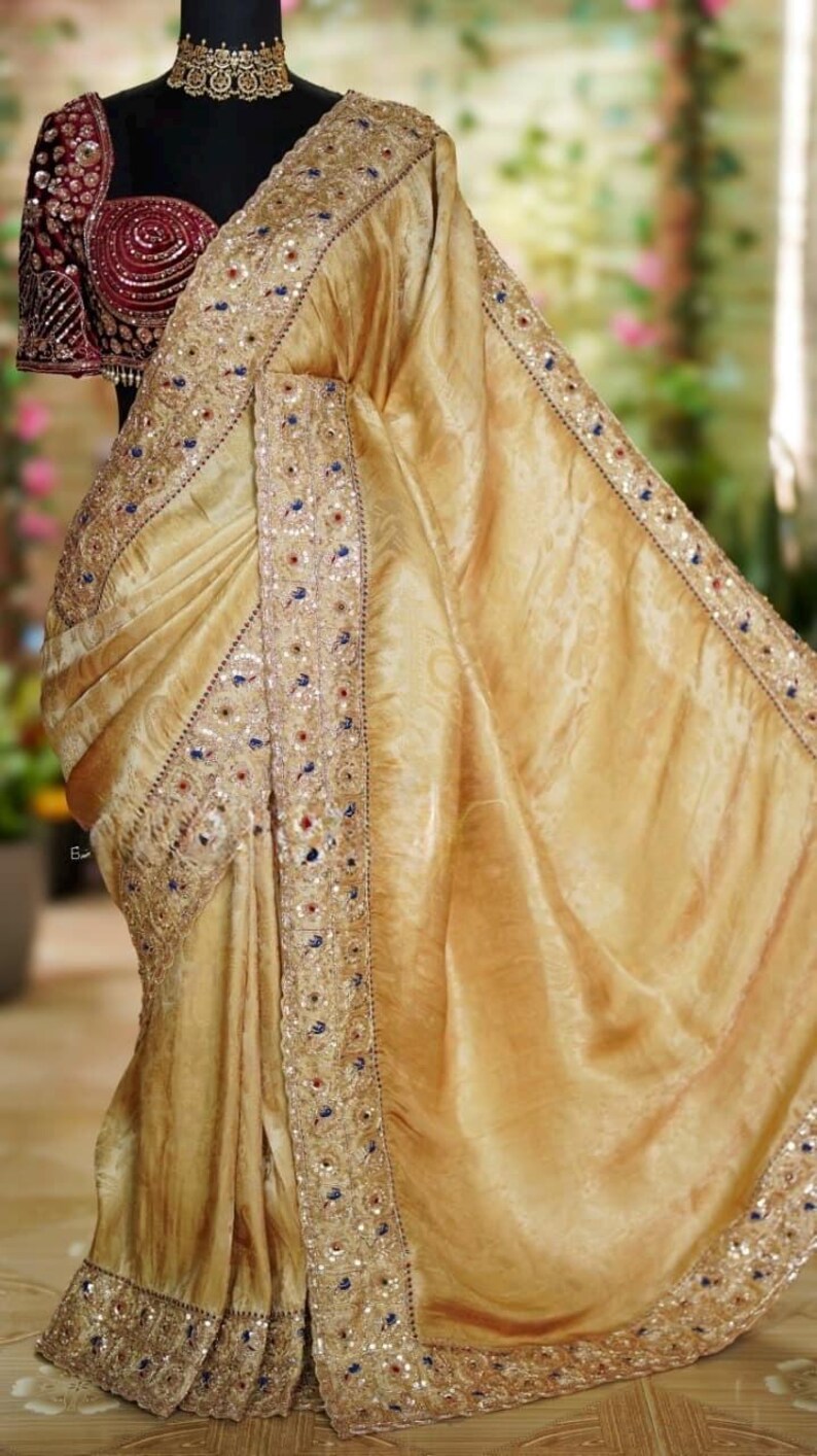 May include: A golden-yellow saree with intricate embroidery and sequin detailing. The border features a pattern of sequins and small embellishments. A maroon blouse with ornate embroidery is visible.