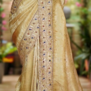 May include: A golden-toned sari with intricate embroidery and embellishments. The border features detailed patterns with sequins and colorful accents. The fabric drapes elegantly, showcasing the rich texture and design of the traditional garment.