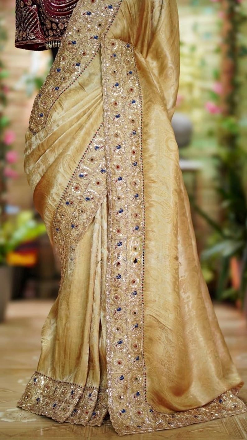 May include: A golden-toned sari with intricate embroidery and embellishments. The border features detailed patterns with sequins and colorful accents. The fabric drapes elegantly, showcasing the rich texture and design of the traditional garment.