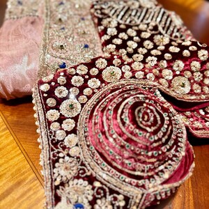 May include: Close-up of ornate, embroidered garments. A pink, patterned fabric with gold and blue embellishments is visible, along with a deep red velvet piece adorned with sequins and beads. The pieces appear to be part of a traditional outfit.