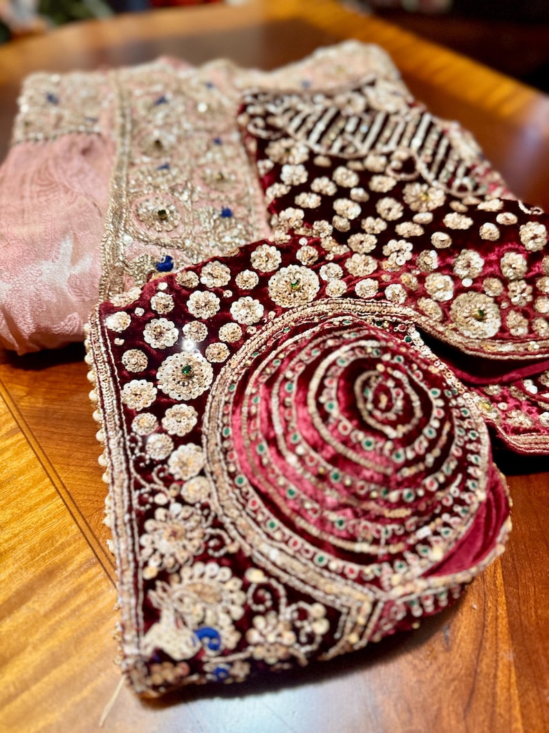May include: Close-up of ornate, embroidered garments. A pink, patterned fabric with gold and blue embellishments is visible, along with a deep red velvet piece adorned with sequins and beads. The pieces appear to be part of a traditional outfit.