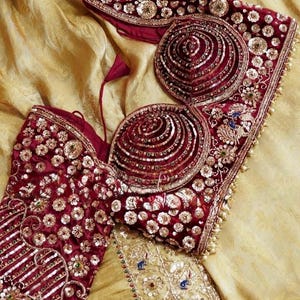 May include: A detailed shot of a maroon and gold embellished bodice and matching top. The garments are heavily decorated with sequins, beads, and embroidery. The background is a golden-yellow fabric, creating a luxurious feel.