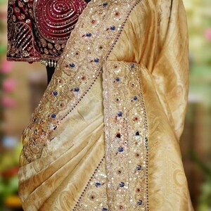 May include: A gold-colored saree with intricate embroidery and embellishments. The border features detailed patterns with sequins and colorful accents. The blouse is a deep burgundy with ornate detailing and a matching gold choker.