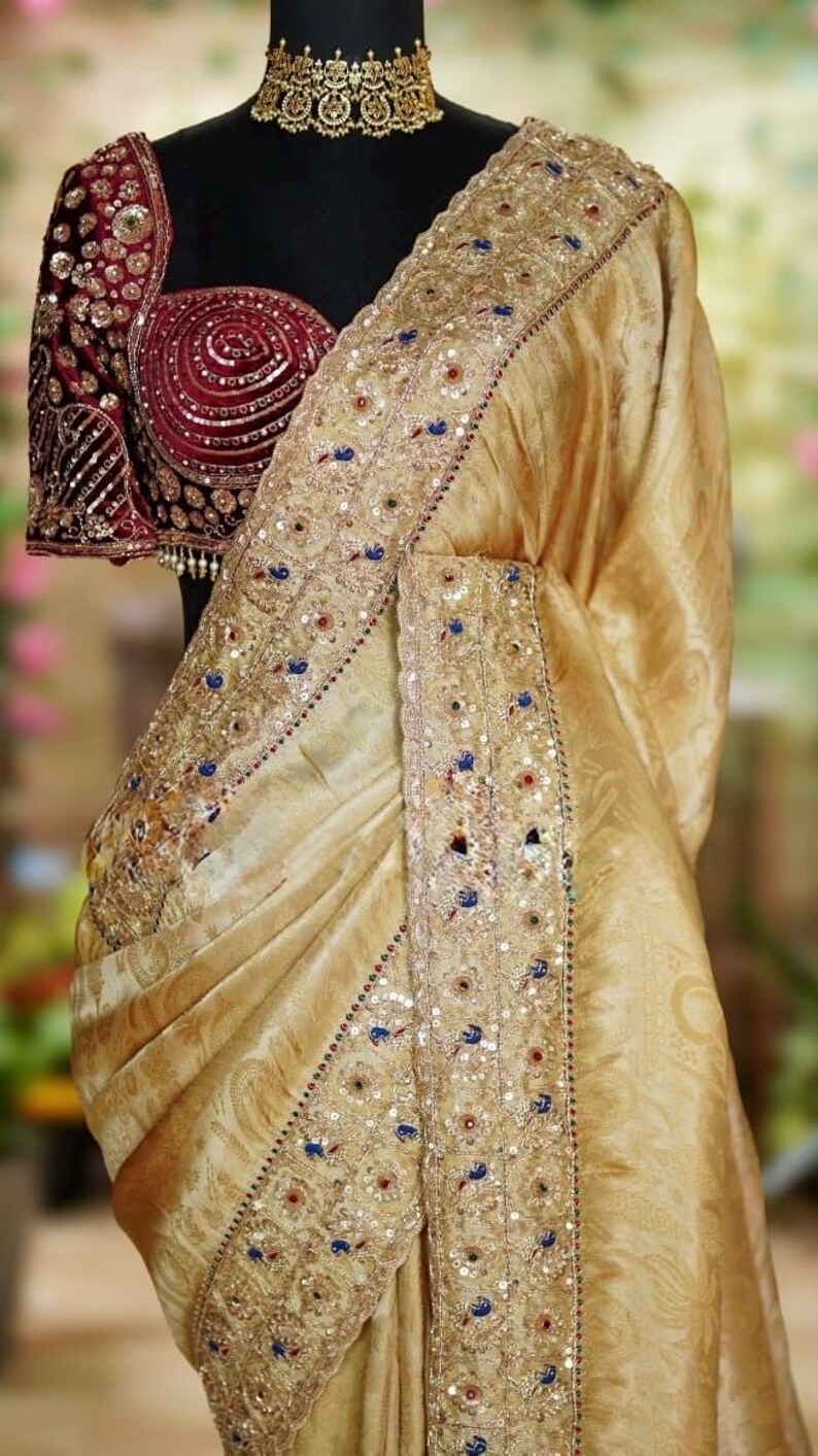May include: A gold-colored saree with intricate embroidery and embellishments. The border features detailed patterns with sequins and colorful accents. The blouse is a deep burgundy with ornate detailing and a matching gold choker.