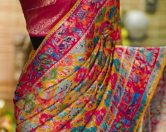 May include: A vibrant, patterned saree in shades of pink, yellow, and blue. The fabric drapes over a mannequin, showcasing intricate designs and a contrasting maroon blouse. A gold-toned necklace adds a touch of elegance.