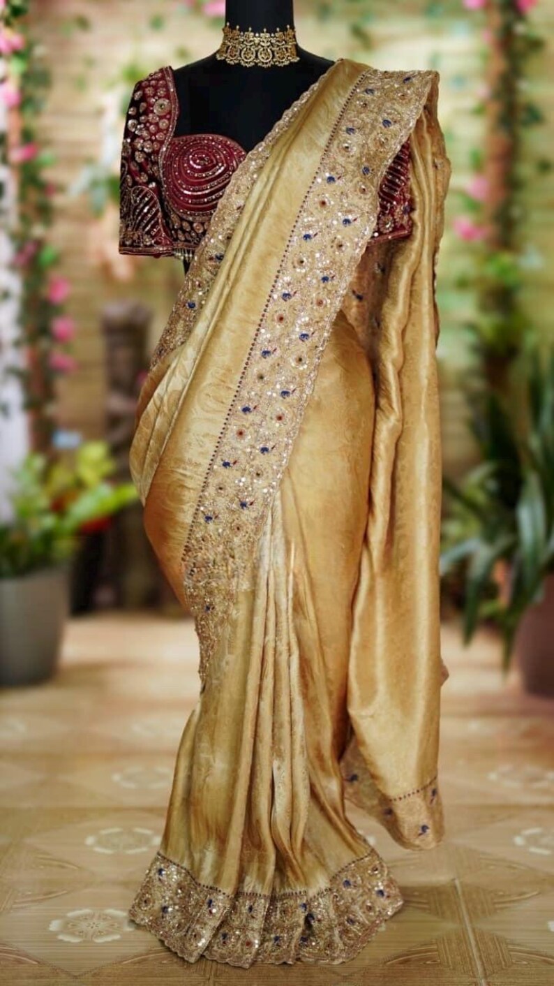 May include: A golden-yellow sari with intricate embroidery and embellishments. The sari features a detailed border with blue and red accents. The blouse is a deep burgundy color with ornate detailing. The garment is draped over a mannequin.