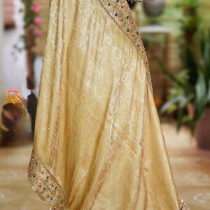 May include: A gold-colored sari with intricate floral patterns and a decorative border. The sari is draped over a mannequin, revealing a maroon blouse with detailed embellishments. The border is adorned with sequins and blue accents.
